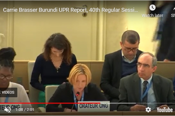 [ai] A woman speaking at a podium during a session, with a report titled 'Burundi UPR Report' in front of her. Two colleagues are seated nearby, and a person is seated in the background, focused on their device.